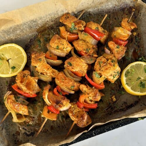 three cooked chicken tikka skewers on baking tray with lemons on side