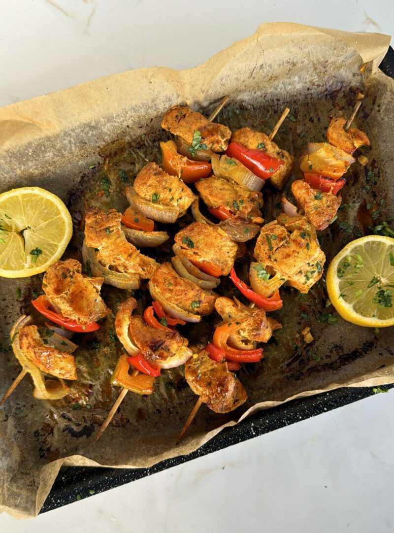 three cooked chicken tikka skewers on baking tray with lemons on side