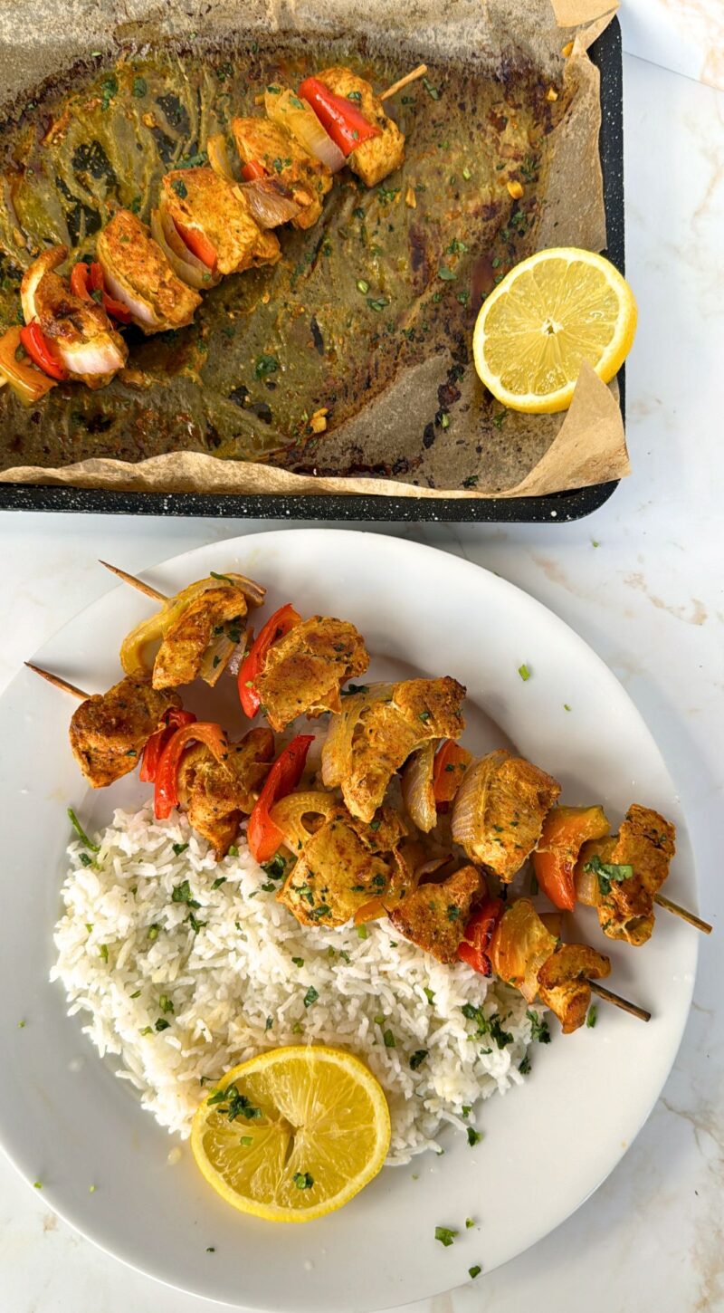 Cooked chicken tikka with rice on a plate and skewer on baking tray with lemons on the side of both